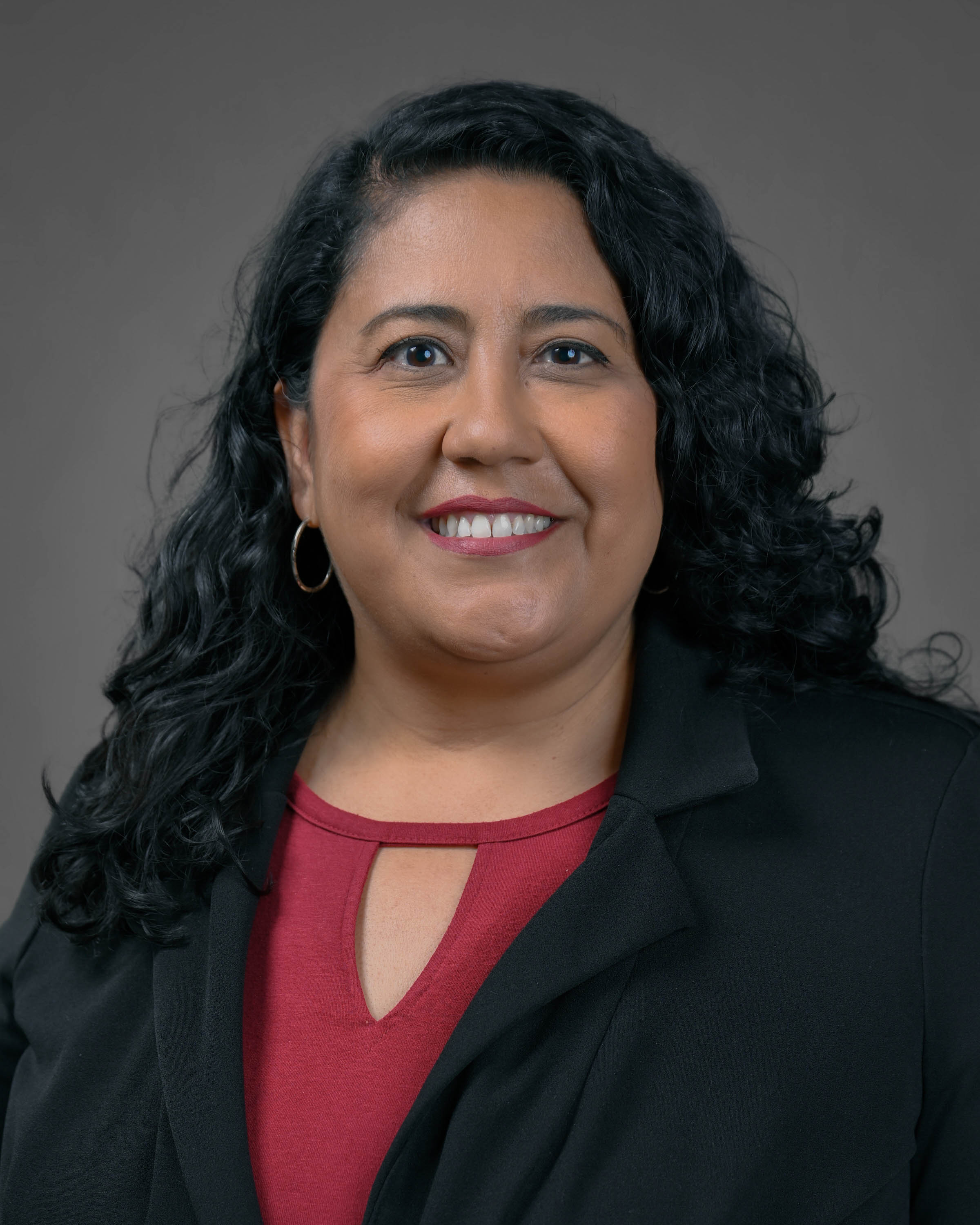 Professional headshot of a smiling woman with curly dark hair, wearing a black blazer and red blouse against a gray studio background.